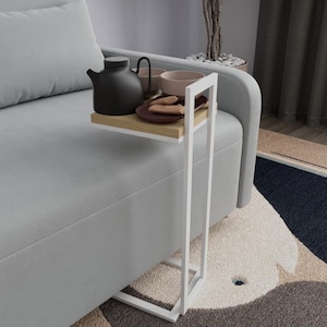 Puede incluir: Una mesa auxiliar de metal blanco con una estantería de madera clara. Una tetera negra, dos tazas y un plato con galletas están en la estantería. La mesa está al lado de un sofá gris.