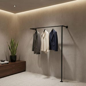May include: A black metal clothes rail displaying three items of clothing: a grey blazer, a white blouse, and a navy blue blazer. The rail is fixed to a grey wall, creating a minimalist aesthetic.