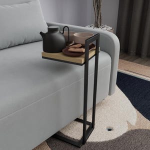 Könnte beinhalten: Ein schwarzer Beistelltisch aus Metall mit einem hellen Holzregal hält eine schwarze Teekanne, zwei Tassen und einen Teller mit Keksen. Der Tisch hat ein rechteckiges Regal und ein schwarzes Metallgestell. Der Tisch steht neben einem grauen Sofa.