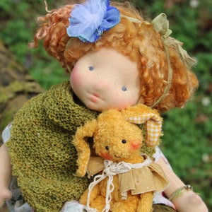 Może przedstawiać: Ręcznie robiona lalka z kręconymi czerwonymi włosami, ubrana w zielony dzianinowy szalik i trzymająca żółtego pluszowego króliczka z biało-żółtą kratkowaną kokardką.