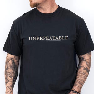 May include: Black crew neck t-shirt with the word "UNREPEATABLE" printed in a light tan color. The text is centered above a thin, horizontal line. The shirt is short-sleeved.