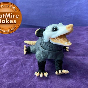 May include: A gray and white opossum toy with a wide open mouth and black eyes. The toy is made of plastic and has a textured surface.