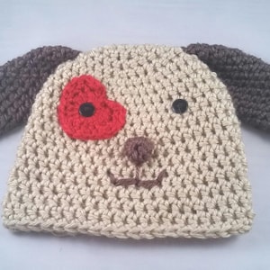 May include: A crocheted dog hat in beige with dark gray ears. The hat features a red heart-shaped patch with a black button, a brown nose, and stitched details for the mouth. A cute and cozy accessory.
