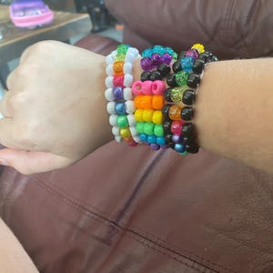 Könnte beinhalten: Eine Sammlung bunter Perlenarmbänder. Die Armbänder weisen eine Vielzahl von Farben auf, darunter Regenbogen, Schwarz, Weiß und Glitzerakzente. Die Armbänder sind an einem Handgelenk gestapelt.