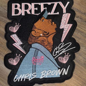 Puede incluir: Parche bordado con el nombre "BREEZY" en rosa, sobre una ilustración de una persona mirando hacia arriba. El parche incluye el texto "CHRIS BROWN" y una firma, con rayos y corazones rosas.