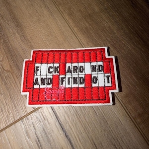 May include: A rectangular embroidered patch featuring a red grid pattern on a white background. The text FUCK AROUND AND FIND OUT is stitched in black capital letters across the center. A small logo with the text The Sparkle Society is visible.
