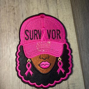 May include: An embroidered patch featuring a person wearing a bright pink baseball cap with the text SURVIVOR and a pink ribbon. The person has dark skin, full pink lips, and black hair. Two pink ribbon earrings hang from the ears of the figure.