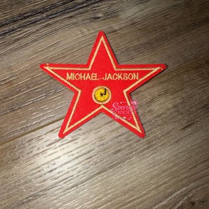 Puede incluir: Parche rojo en forma de estrella con ribete dorado y el nombre "MICHAEL JACKSON" bordado. Una pequeña carita sonriente amarilla está en el centro. El parche está sobre una superficie de madera.