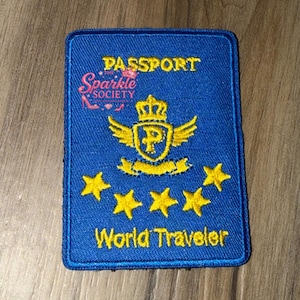 Peut inclure: Un écusson rectangulaire bleu avec le mot "PASSPORT" en haut et "World Traveler" en bas. L'écusson présente un emblème doré avec des ailes, une couronne et la lettre "P", ainsi que cinq étoiles dorées.