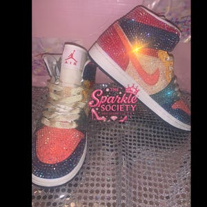Puede incluir: Un par de zapatillas altas, muy adornadas con brillantes pedrer&iacute;a en una vibrante gama de colores. Los zapatos tienen una suela blanca, un logotipo de Nike y el texto "AIR" en la leng&uuml;eta. Los zapatos forman parte de la colecci&oacute;n "The Sparkle Society".
