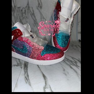 Puede incluir: Un par de zapatillas personalizadas con suela y empeine blancos. La puntera, el tal&oacute;n y los paneles laterales est&aacute;n cubiertos de pedrer&iacute;a rosa, morada y turquesa. El logo de Nike es turquesa. El texto "The Sparkle Society" es visible.