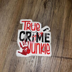 Peut inclure: Écusson brodé avec les mots "True Crime Junkie" en rouge et noir, avec un dessin d'empreinte de main, de couteau et de loupe. L'écusson a une bordure blanche.