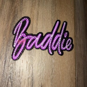 May include: A decorative patch featuring the word Baddie in a cursive script font. The lettering is outlined in bright purple and filled with clear, sparkling rhinestones, all set against a solid black background with a glittery texture.