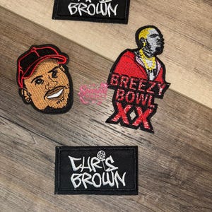 Puede incluir: Parches bordados con Chris Brown. Un parche tiene el texto "CHRIS BROWN" en blanco sobre un rectángulo negro. Otro parche es un retrato de Chris Brown con una gorra roja. Un tercer parche tiene el texto "BREEZY BOWL XX".