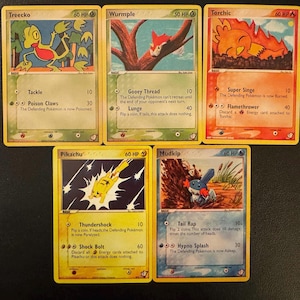 May include: Five Pokémon trading cards featuring Treecko, Wurmple, Torchic, Pikachu, and Mudkip. Each card has a unique illustration and attack descriptions. The cards are yellow with various colors and designs.