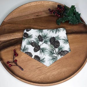 May include: A white bandana with a pine cone and evergreen branch pattern, displayed on a wooden tray. The bandana features a repeating design of dark brown pine cones and green pine needles. Decorative red berries and green leaves are also present.