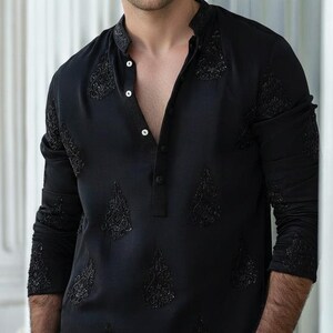May include: Black long-sleeve shirt with intricate embroidered detailing. The shirt features a mandarin collar and a button-down placket. The text "AM SHIFACRAFTDESIGN" is displayed at the top right corner. The shirt is made of a soft, comfortable material.