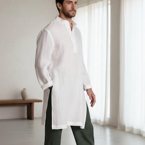 Puede incluir: Una túnica de lino blanco con cuello mandarín y mangas largas, combinada con pantalones de lino verde oscuro. El conjunto forma parte de la colección Shifacraft. El modelo está descalzo en un entorno luminoso y minimalista.