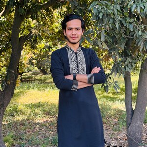 May include: A black tunic with intricate silver and gold embroidery around the collar and cuffs. The tunic is worn by a person standing outdoors with arms crossed. The text "AM SHIFACRAFT" is visible at the bottom of the image.