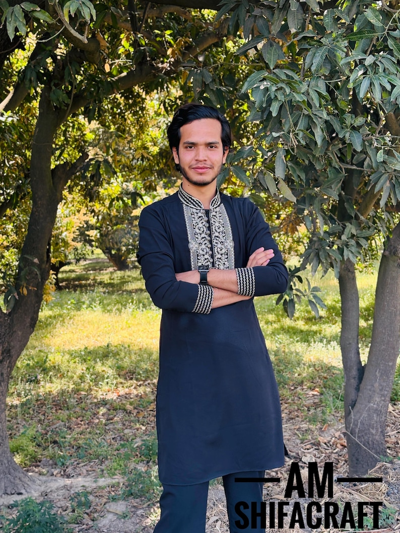 May include: A black tunic with intricate silver and gold embroidery around the collar and cuffs. The tunic is worn by a person standing outdoors with arms crossed. The text "AM SHIFACRAFT" is visible at the bottom of the image.