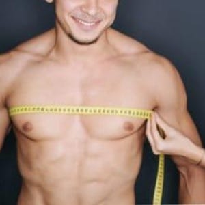 May include: A person's chest is being measured with a yellow measuring tape. The tape is held horizontally across the chest. The person is smiling. The background is a dark gray color.