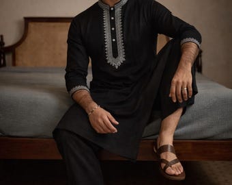 Op de afbeelding: Zwart kurta-pyjamaset met ingewikkelde witte borduursels op de kraag en manchetten. De outfit wordt gecombineerd met bruine sandalen. De afbeelding bevat de tekst "AM SHIFACRAFTDESIGN".