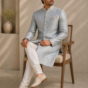 Pu&ograve; includere: Una giacca lunga fantasia blu chiaro e oro con bottoni abbinati, abbinata a pantaloni bianchi e mocassini color crema. L'outfit &egrave; indossato da una persona seduta su una sedia di legno. &Egrave; visibile un fazzoletto da taschino. Il testo "AM SHIFACRAFT" &egrave; in basso.