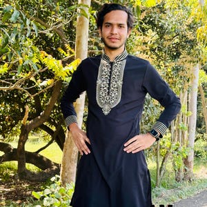 May include: A black tunic with intricate white and gold embroidery around the collar and cuffs. The long-sleeved tunic is paired with matching black pants. The image also includes the text "AM-SHIFACRAFT".