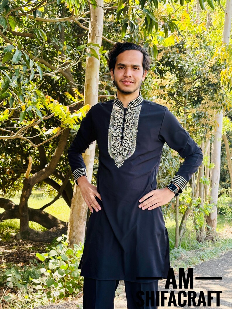 May include: A black tunic with intricate white and gold embroidery around the collar and cuffs. The long-sleeved tunic is paired with matching black pants. The image also includes the text "AM-SHIFACRAFT".