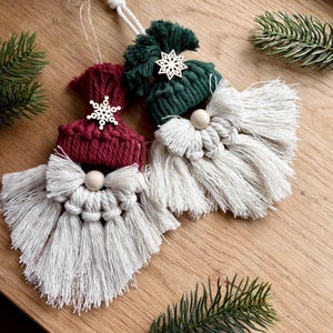 Könnte beinhalten: Nahaufnahme von zwei handgefertigten Weihnachtsschmuckstücken. Eines ist weinrot, das andere grün. Beide haben einen Makramee-Bart, eine Holzkugel-Nase und eine Schneeflocke oben. Der Schmuck liegt auf einer Holzoberfläche mit Tannennadeln.