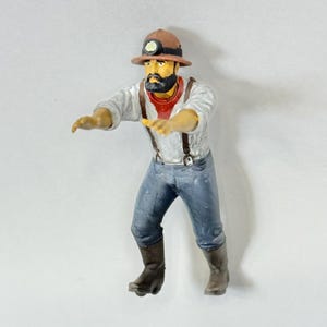 May include: A figurine of a miner, wearing a brown hard hat with a lamp, a white shirt, blue jeans, brown boots, and suspenders. The miner has a black beard and a red neckerchief. The figurine is posed with arms outstretched.