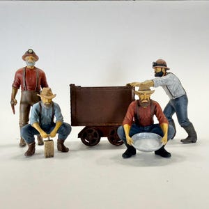 May include: A collection of miniature figurines depicting gold miners. The figures are dressed in work clothes, with hats and tools. One figure is pushing a rusty brown cart, while another holds a pan. The scene evokes the spirit of the gold rush era.