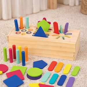 Puede incluir: Un juguete de clasificación de formas de madera con una variedad de formas geométricas coloridas y clavijas. El clasificador tiene un acabado de madera natural y presenta agujeros para las formas y las clavijas. Las formas incluyen círculos, cuadrados, triángulos y otras formas en varios colores.