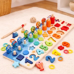 Può includere: Giocattolo educativo in legno con forme, numeri e pezzi a forma di pesce colorati. Il giocattolo include pioli con anelli impilati e pezzi a forma di pesce. La base è dipinta con onde blu e bianche. È inclusa anche una matita di legno.