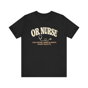 May include: A black short-sleeve t-shirt featuring the cream-colored text OR NURSE in an arched font. Below the text is a graphic of a stethoscope forming a heart shape, followed by the text COLD ROOMS, WARM BLANKETS, SHARP OBJECTS.