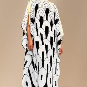 May include: A loose-fitting kaftan dress featuring a bold black and white graphic pattern of repeating arched shapes. The garment has wide, open sleeves and a cream-colored upper section, paired with black dress shoes to complete the ensemble.