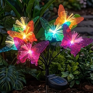 Pode incluir: Uma estaca de jardim movida a energia solar com seis luzes de borboleta coloridas. As borboletas são em vários tons de rosa, laranja, verde e azul, com luzes de fibra ótica. A estaca tem uma base preta e um pequeno painel solar.