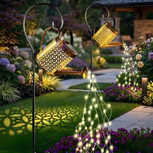 Puede incluir: Dos luces decorativas solares con forma de regadera con acabado bronce, colgadas de ganchos de pastor de metal negro. Las regaderas tienen un diseño estampado que proyecta patrones de luz en el suelo. Cadenas de luces LED blancas cálidas caen de las boquillas.