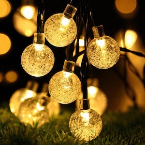 May include: String of warm white globe lights. Each clear globe has a textured, speckled appearance and is lit from within. The lights are strung on a black wire, with a few globes in focus against a blurred background of additional lights.