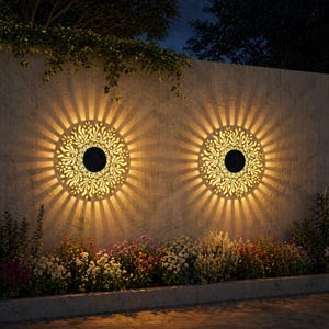 Op de afbeelding: Twee decoratieve buitenmuurlampen met ingewikkelde bloempatronen werpen een warme, radiale gloed. De lampen zijn gemonteerd op een getextureerde muur boven een bloembed met kleurrijke bloemen. De lampen hebben een zwart midden.