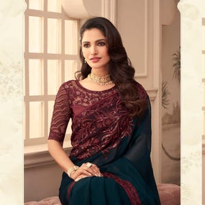 May include: A woman wearing a teal saree with a burgundy embroidered bodice and sleeves. The saree features intricate sequin detailing and a burgundy border. The image includes the text "TFH THE FASHION HUB".