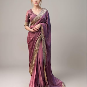 May include: A mauve saree with gold embroidered borders and a matching short-sleeved blouse. The fabric has a subtle sheen, and the saree drapes elegantly. The design features intricate gold detailing along the edges.