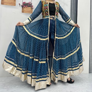 May include: A teal-colored, long, flowing dress with gold trim and a matching embroidered jacket. The jacket has colorful embroidery and a black top underneath. The dress has a striped pattern and is floor-length.
