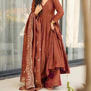 May include: A rust-coloured, long-sleeved dress with a V-neck and flowing skirt. The dress is paired with a matching shawl featuring a white and gold floral pattern. The outfit appears to be made of a textured fabric.