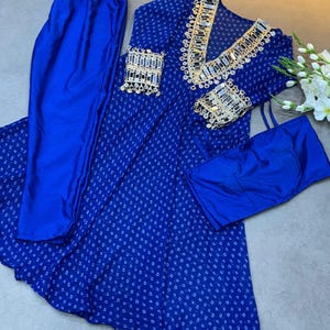 Puede incluir: Un conjunto azul de dos piezas con una parte superior estampada y pantalones a juego. La parte superior presenta un cuello en V y adornos decorativos alrededor del escote y los puños. La tela tiene un sutil patrón de pequeñas formas blancas.
