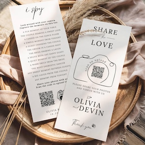 Könnte beinhalten: Zwei weiße Hochzeitskarten mit schwarzem Text, eine mit "i spy" und einer Liste von Aufforderungen, die andere mit "Share the Love" und einer Kameragrafik. Beide Karten haben QR-Codes und Namen. Die Karten liegen auf einem Holztablett.