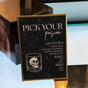 May include: A rectangular sign in a gold frame with a black background featuring the text PICK YOUR posion, a list of cocktails, wine, and beer, and a white line drawing of a human skull inside a glass tumbler, set against a subtle ghost pattern.