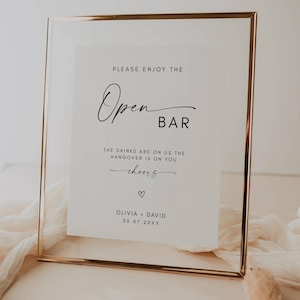 Könnte beinhalten: Ein rechteckiges Schild in einem goldenen Rahmen mit weißem Hintergrund. Der Text in Schwarz lautet: „PLEASE ENJOY THE Open BAR“. Darunter steht „THE DRINKS ARE ON US THE HANGOVER IS ON YOU cheers“ mit einem Herz, Namen und Datum.