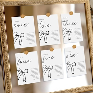 Puede incluir: Una exhibición enmarcada de seis tarjetas de mesa blancas, cada una con un número único en escritura elegante: uno, dos, tres, cuatro, cinco y seis. Cada tarjeta presenta una ilustración de lazo negro y nombres de invitados.