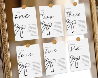 May include: A framed display of six white table cards, each with a unique number in elegant script: one, two, three, four, five, and six. Each card features a black bow illustration and guest names. The cards are affixed with gold wax seals.
